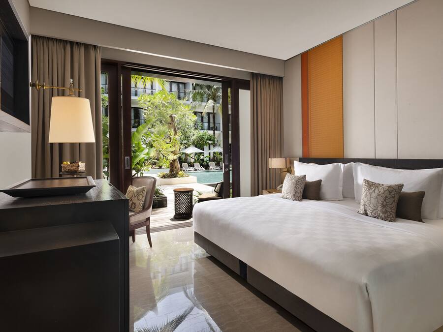A modern hotel room with a large bed, a desk, and a view of the outdoor pool and greenery.