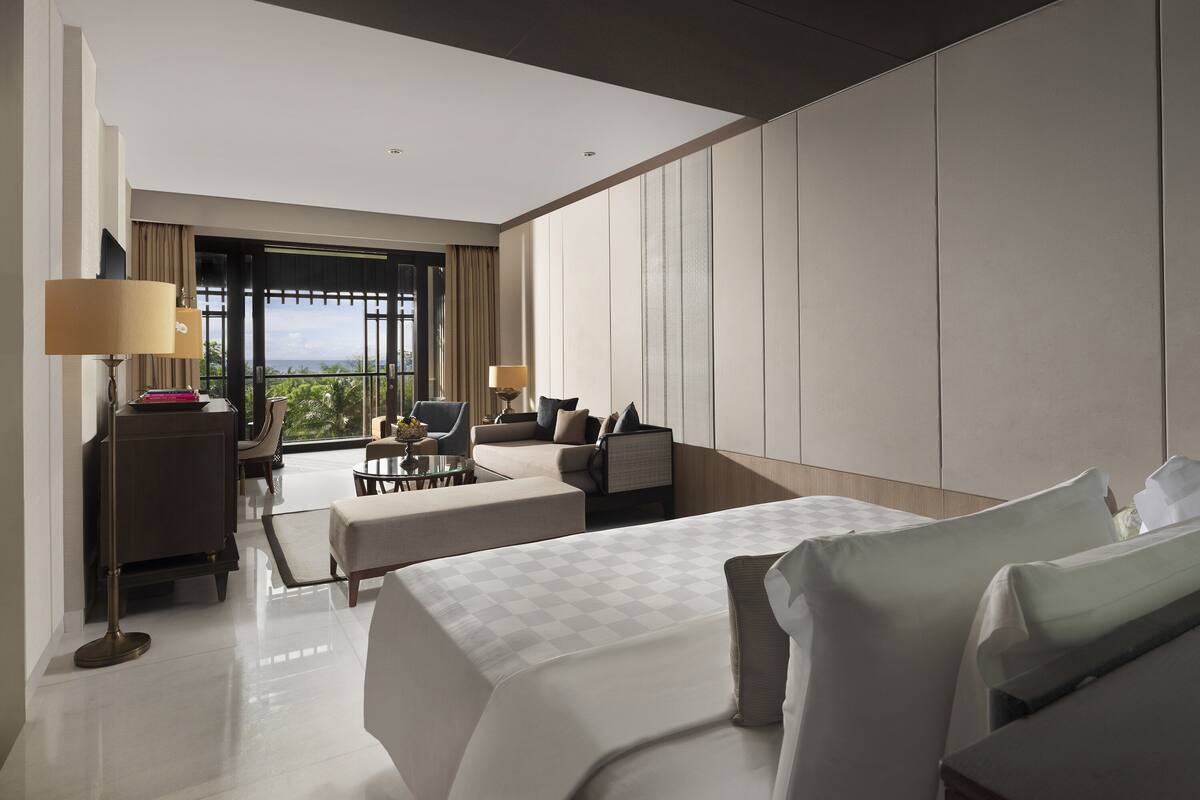 A modern hotel room with a large bed, a seating area with a sofa and chairs, a desk, and a view of the outdoors.