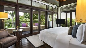 A bedroom with a large bed, a sofa, a small table, and a TV. There are large windows with a view of greenery outside.