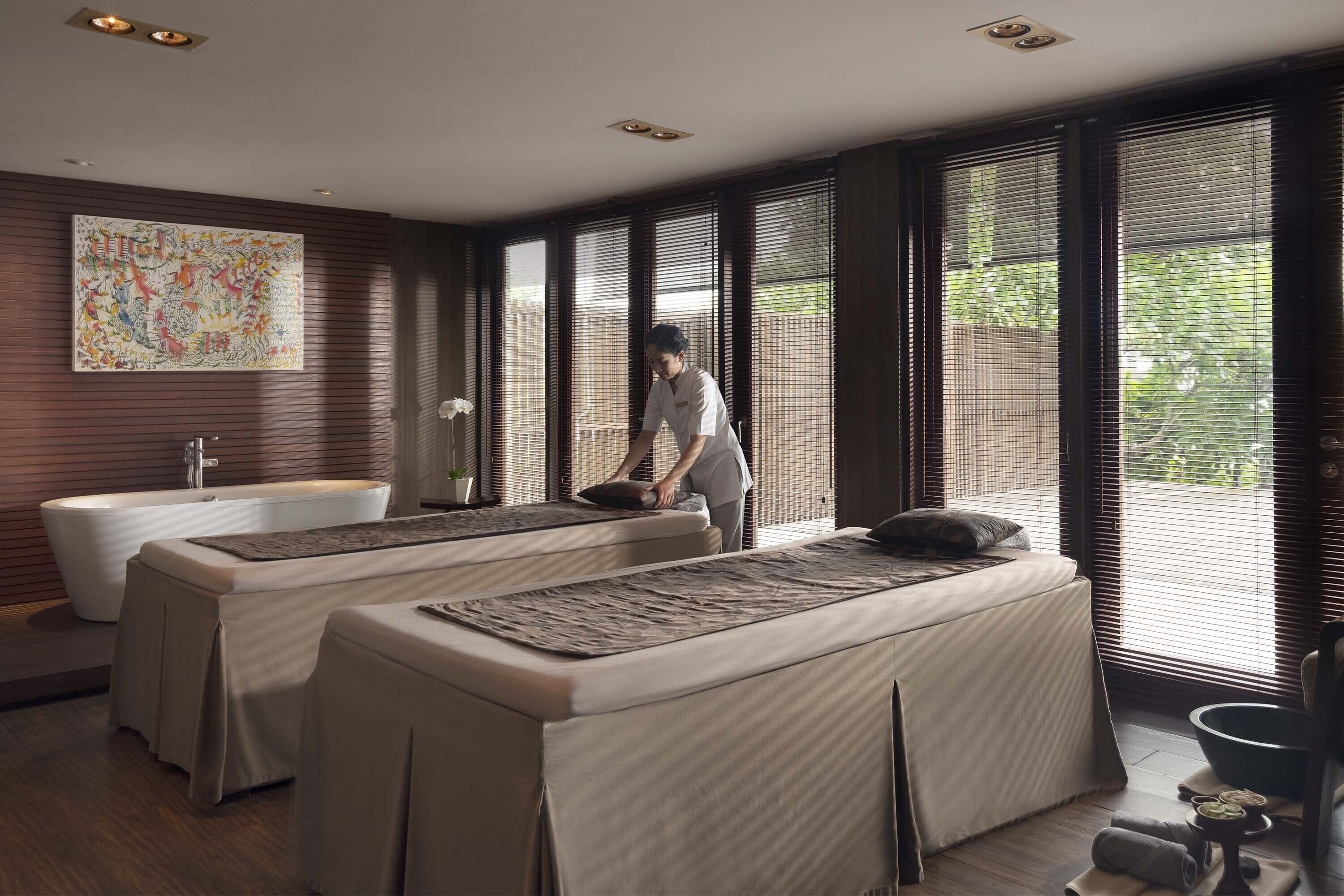 A spa room with two massage tables, a bathtub, and a large window with blinds.