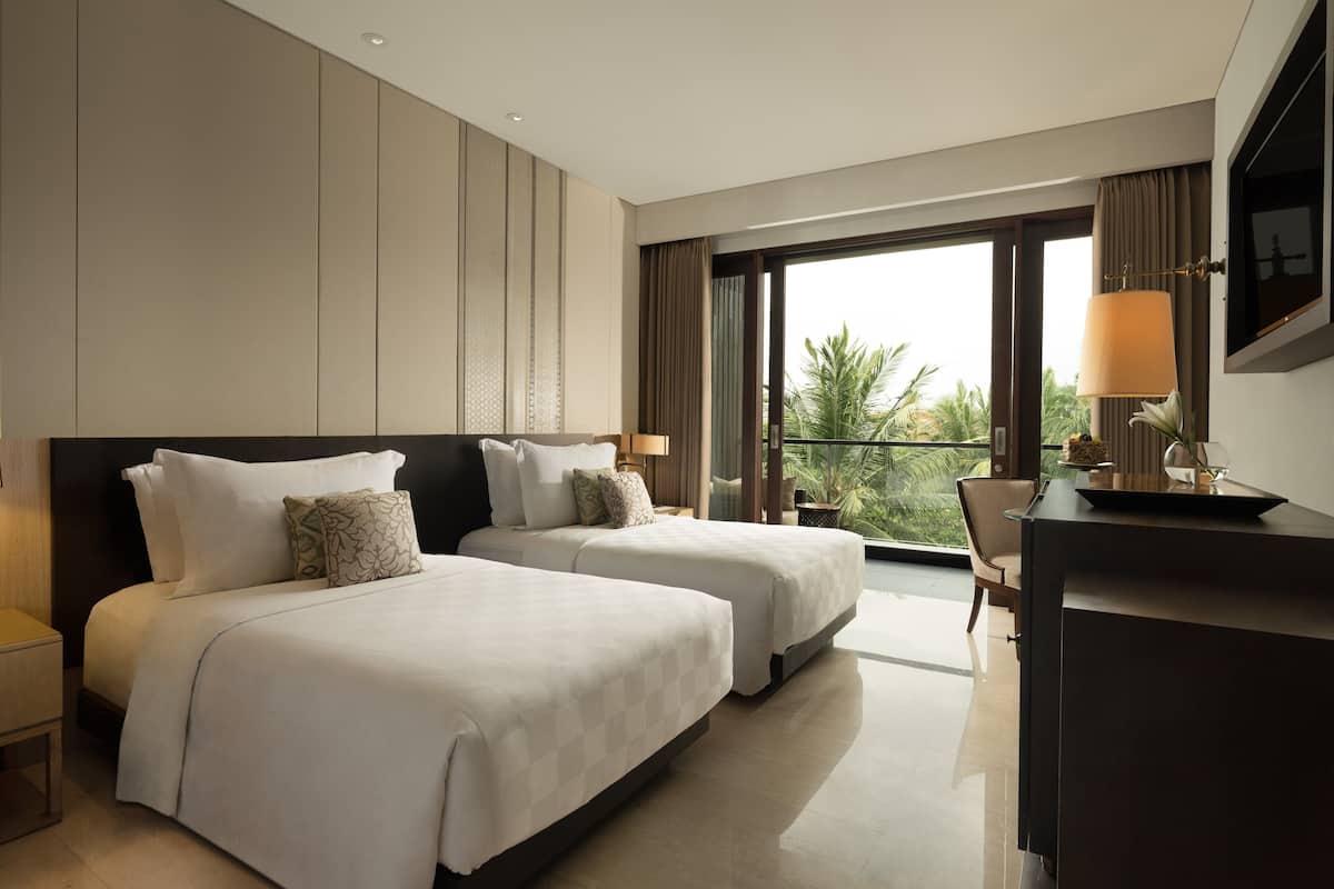 A hotel room with two beds, a desk, a chair, a TV, and a balcony with a view of palm trees.