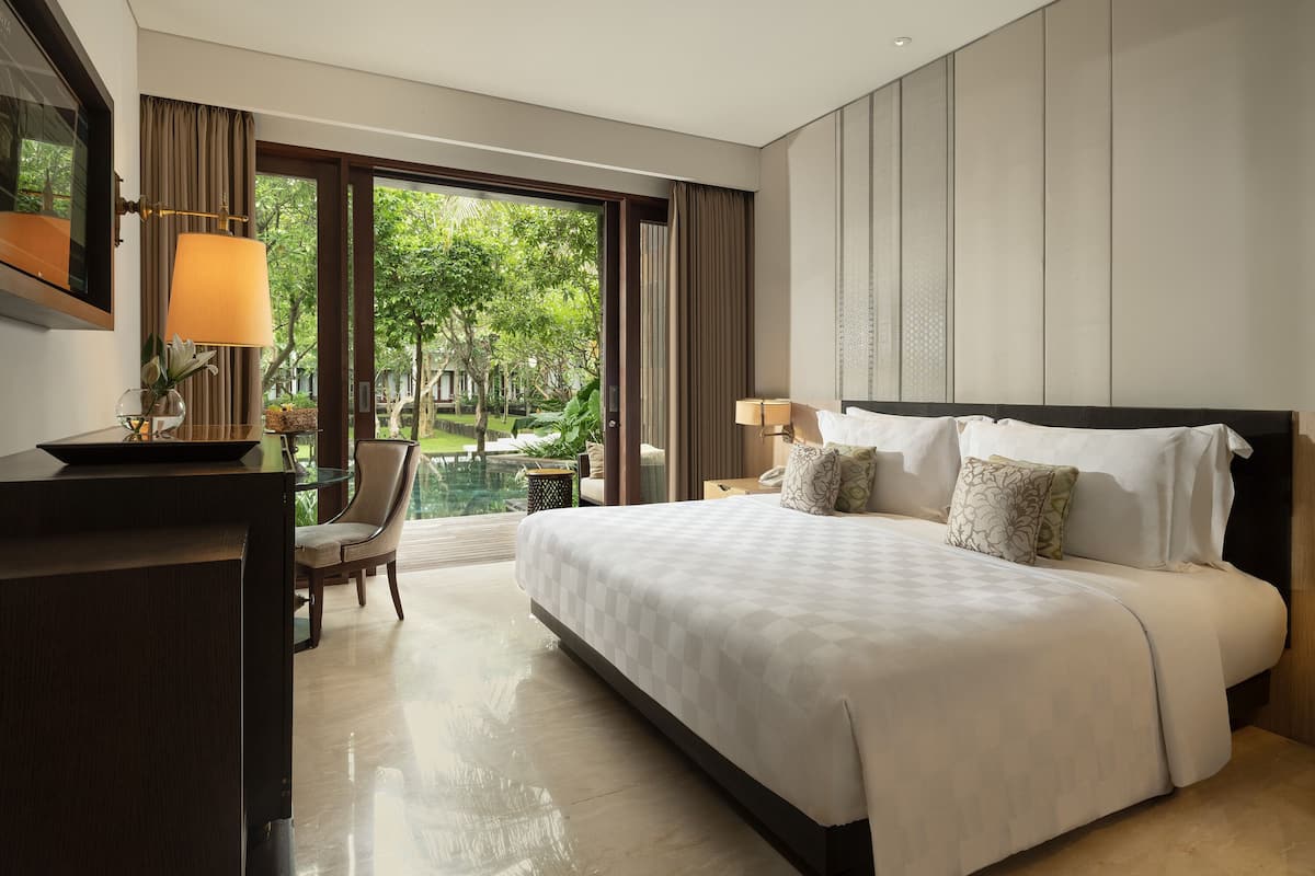 A hotel room with a large bed, a desk, a chair, and a view of a garden through the window.