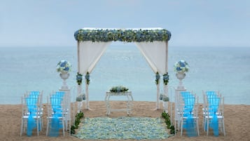 A beach wedding setup with a white arch, blue chairs, and a floral arrangement on the sand.