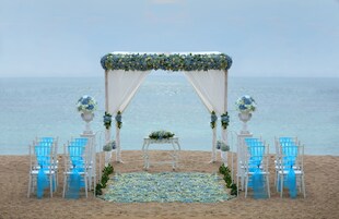A beach wedding setup with a white arch, blue chairs, and a floral arrangement on the sand.
