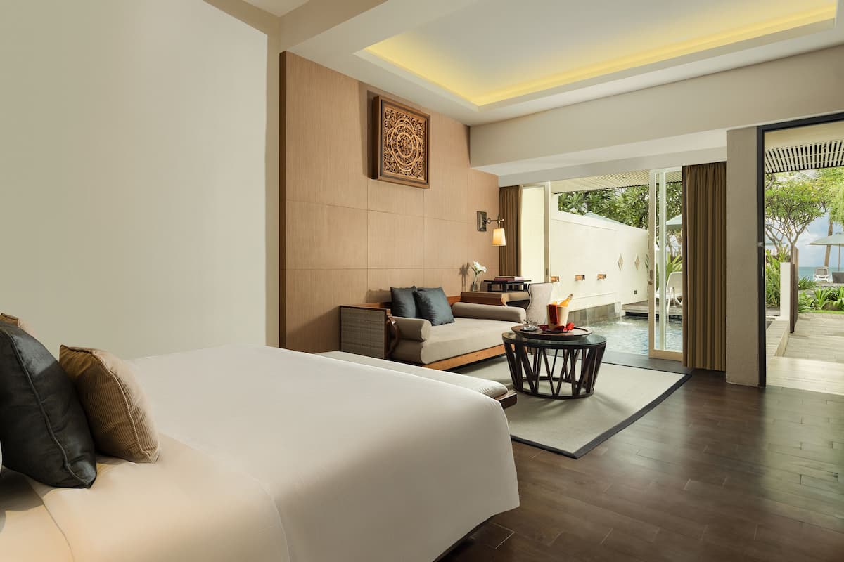 A modern hotel room with a large bed, a sofa, a small table, and a view of the outdoors.
