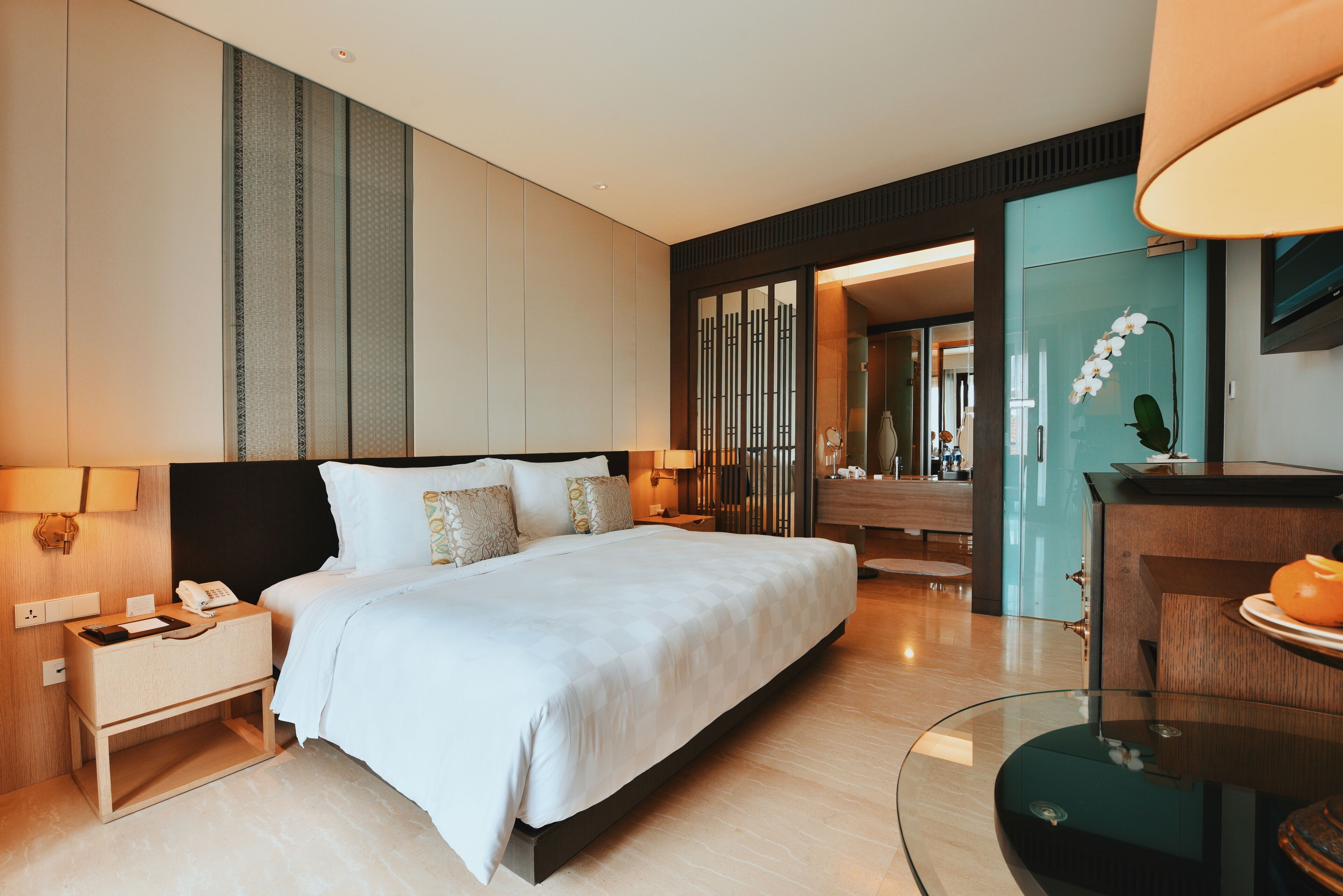 A modern hotel room with a large bed, bedside tables, a glass coffee table, and a bathroom visible through an open door.