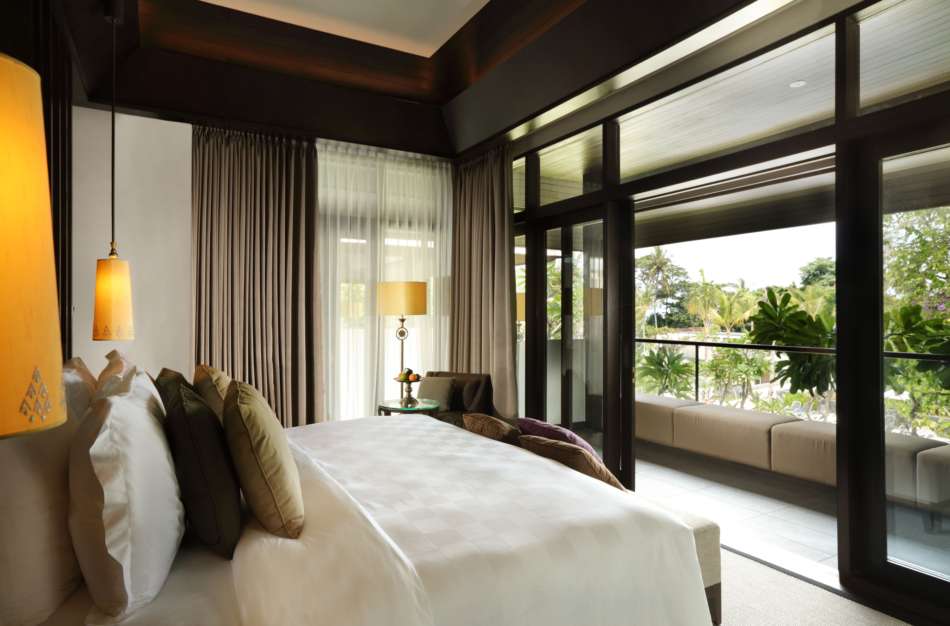 A spacious bedroom with a large bed, a nightstand, a lamp, and a view of the outdoors through large windows.
