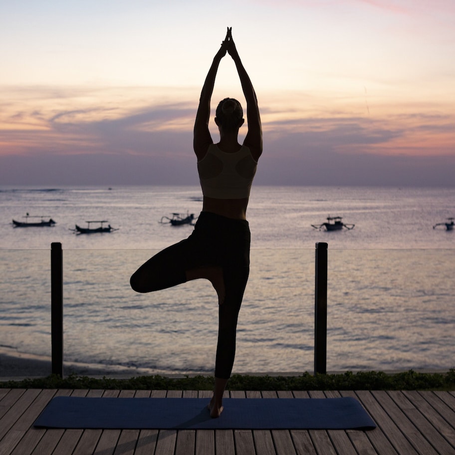 A person performing a yoga pose on a mat with a view of the sea and boats in the background.
