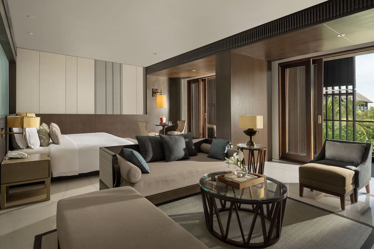 A modern hotel room with a bed, sofa, armchair, and a glass coffee table.