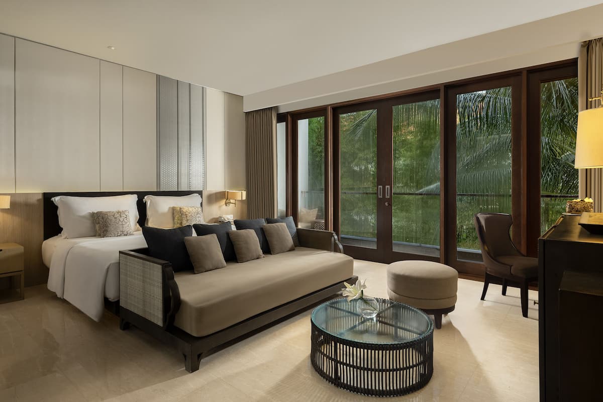 A modern hotel room with a large bed, a sofa, a glass coffee table, and a view of greenery through large windows.