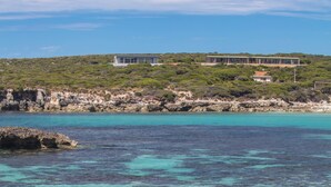 A coastal view with clear turquoise waters, a sandy beach, and a building with large windows overlooking the sea.