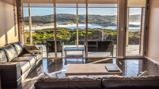 A modern living room with a leather sofa, a coffee table, and a view of a coastal landscape through large glass doors.