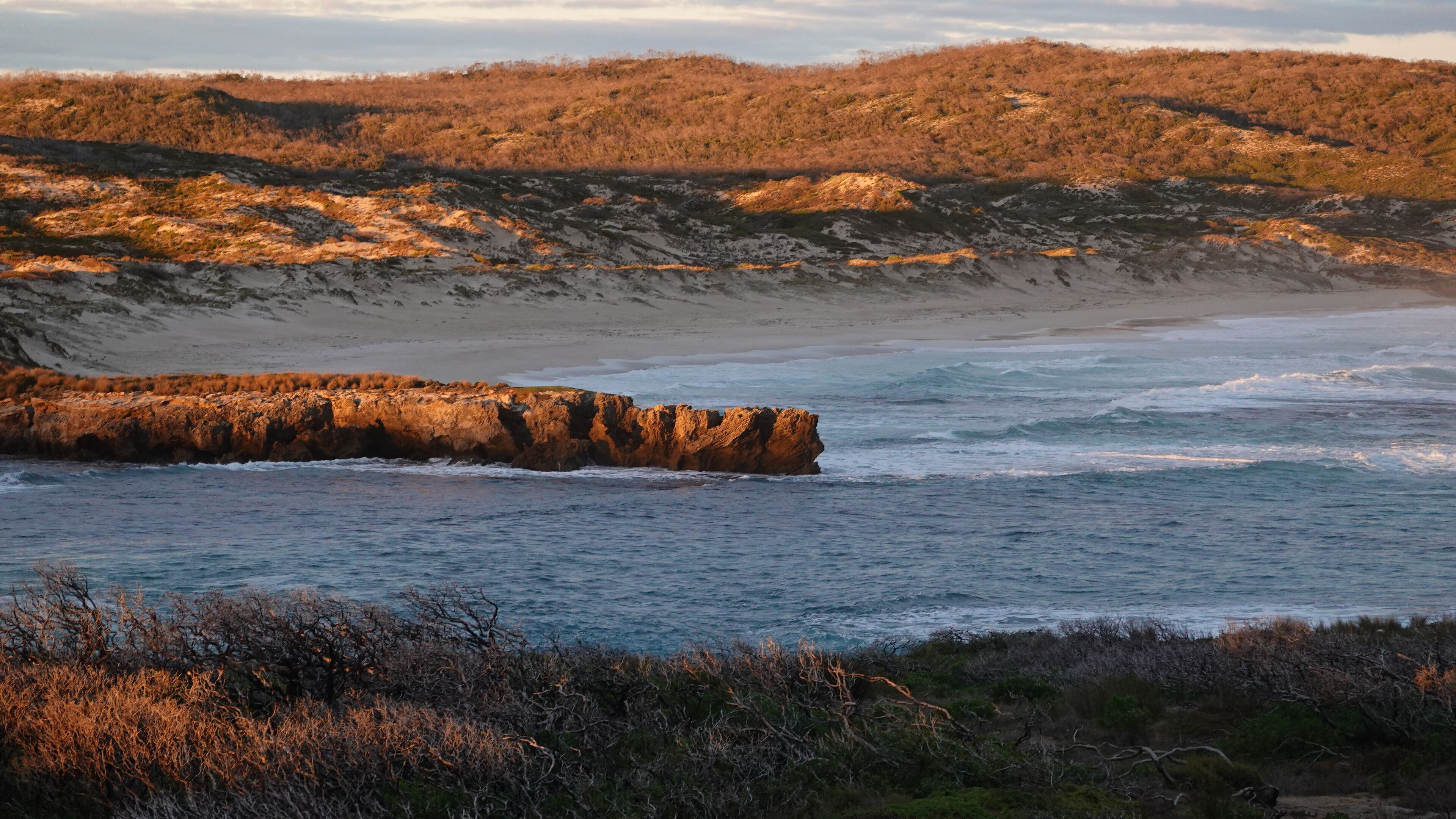Un paysage côtier caractérisé par une formation rocheuse imposante près du rivage, une plage de sable et un arrière-plan vallonné.