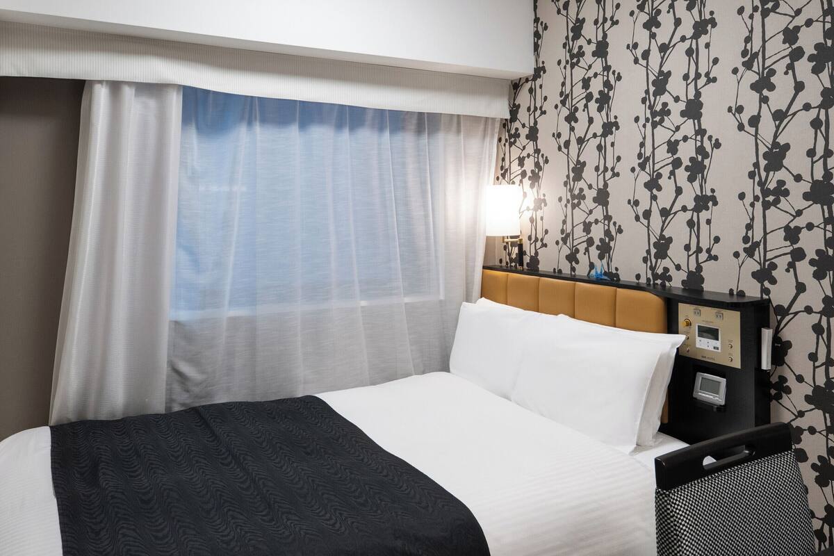 A hotel room with a bed, a nightstand, a lamp, and a wall with a floral pattern.