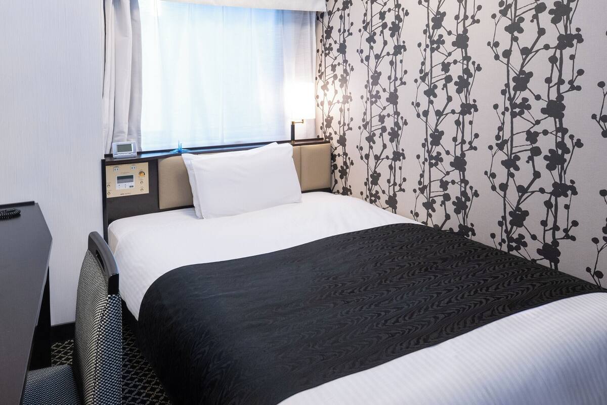 A hotel room with a bed, a desk, a chair, a nightstand, and a wall with a floral pattern.