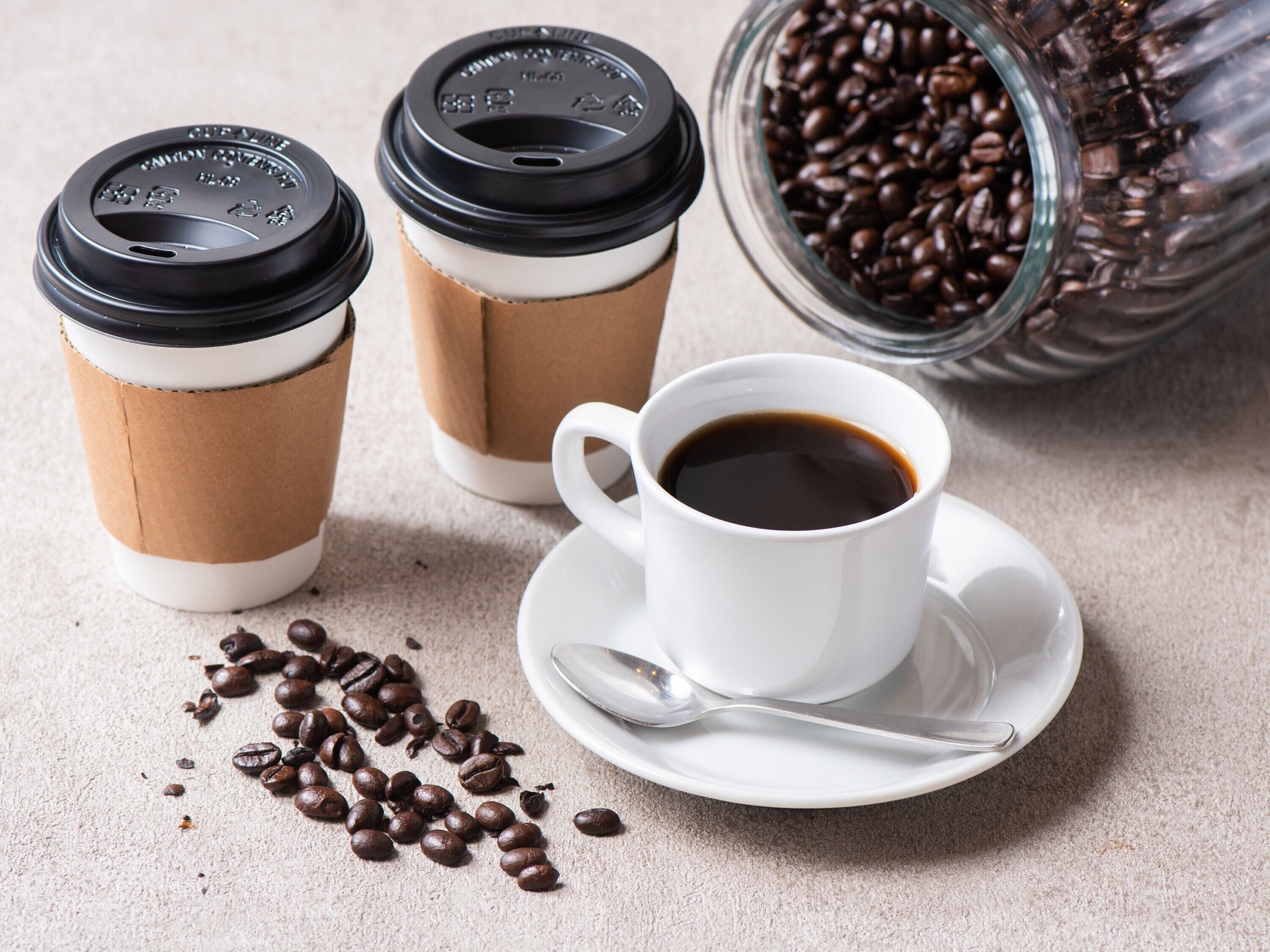 Two disposable coffee cups with lids, a white ceramic cup with a saucer and spoon, and scattered coffee beans on a textured surface.