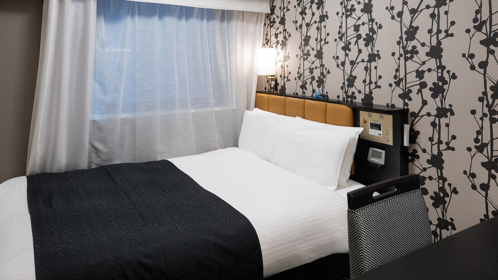 A hotel room with a bed, a nightstand, a lamp, and a wall with a floral pattern.