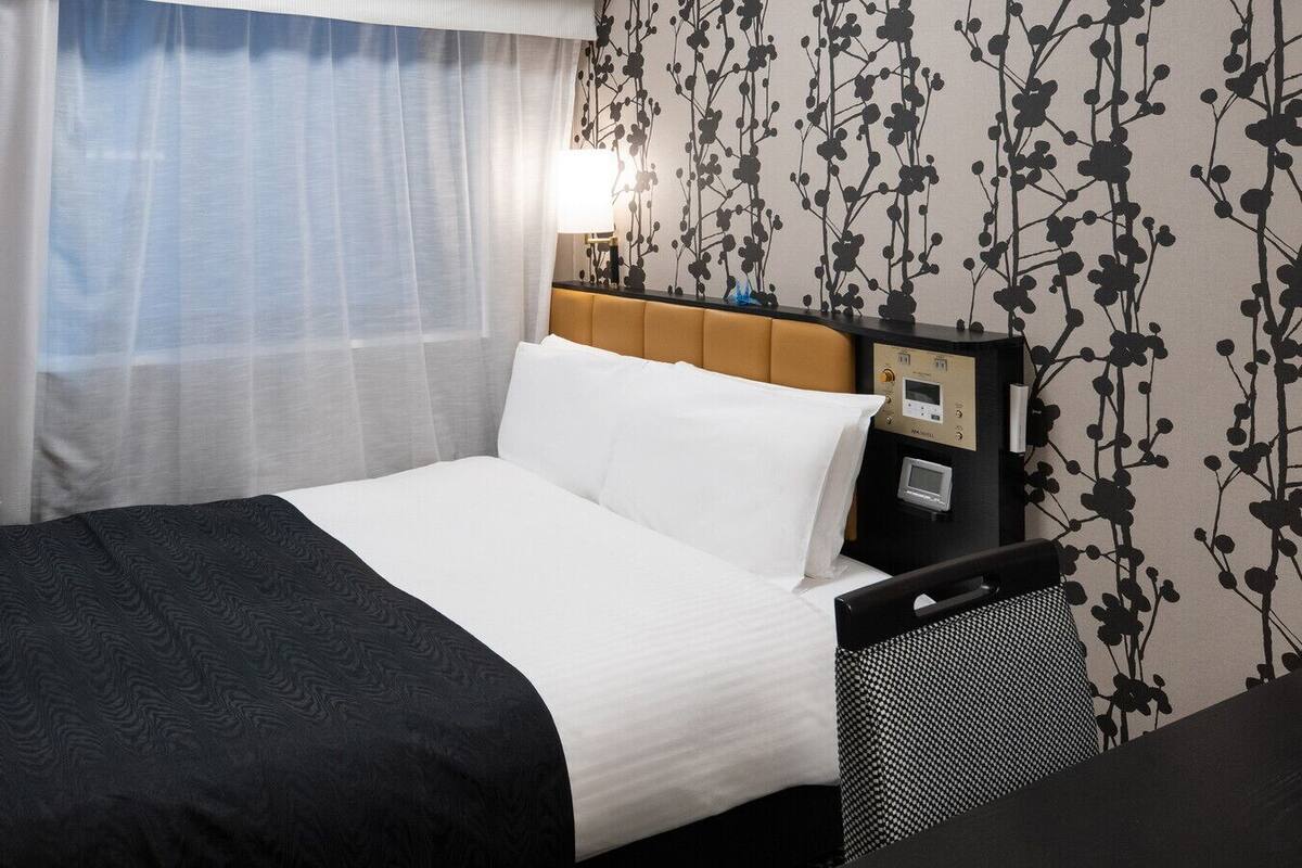 A hotel room with a bed, a nightstand, a lamp, and a wall with a floral pattern.