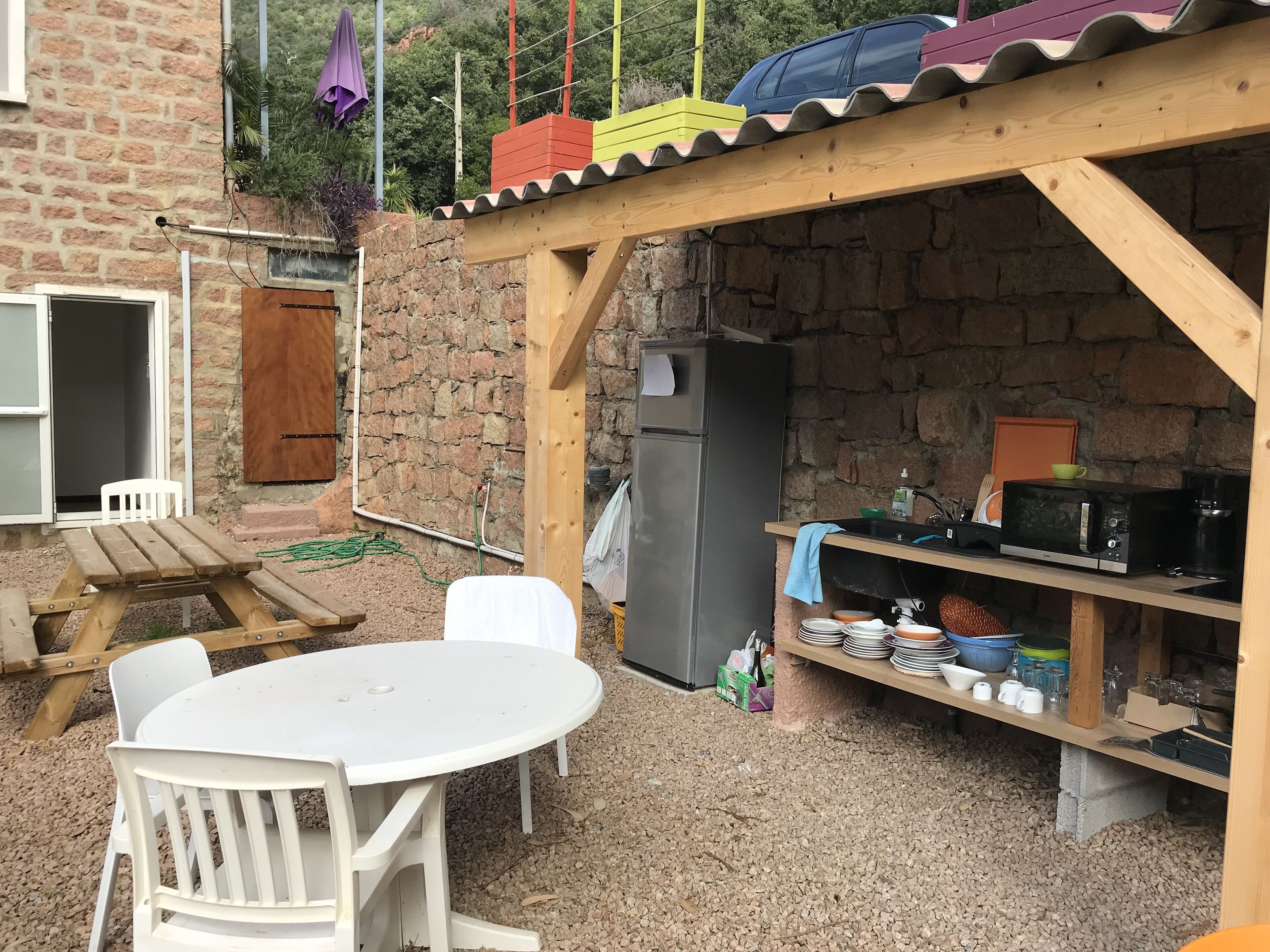 A rustic outdoor kitchen with a wooden table and chairs, a refrigerator, and a shelf with kitchenware.