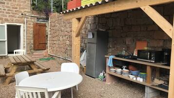 A rustic outdoor kitchen with a wooden table and chairs, a refrigerator, and a shelf with kitchenware.