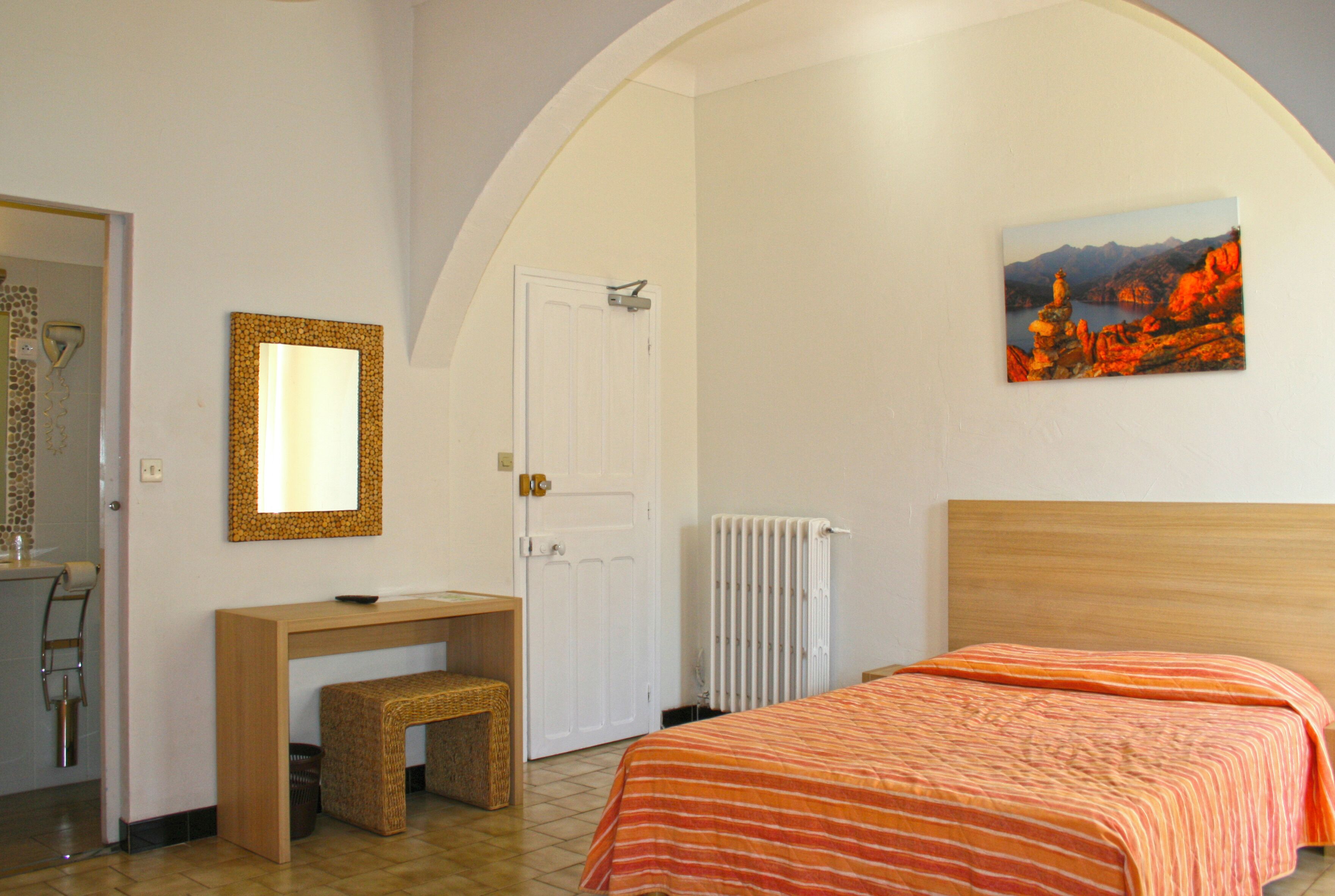A bedroom with a bed, a desk, a chair, a mirror, and a painting on the wall.
