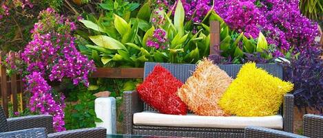 A wicker outdoor seating area with cushions, surrounded by lush greenery and vibrant purple flowers.