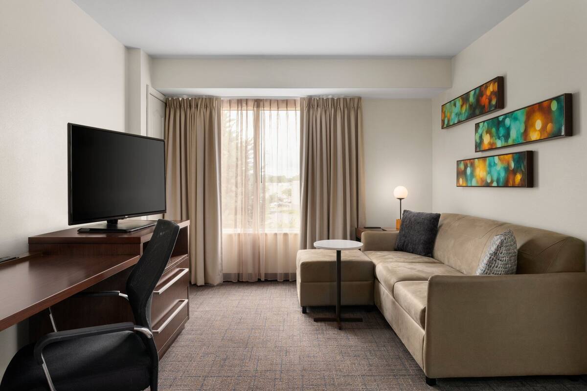 Ein Hotelzimmer mit Flachbildfernseher, Schreibtisch, Sofa und einem kleinen Tisch. An der Wand hängen drei Gemälde und es gibt ein Fenster mit Vorhängen.