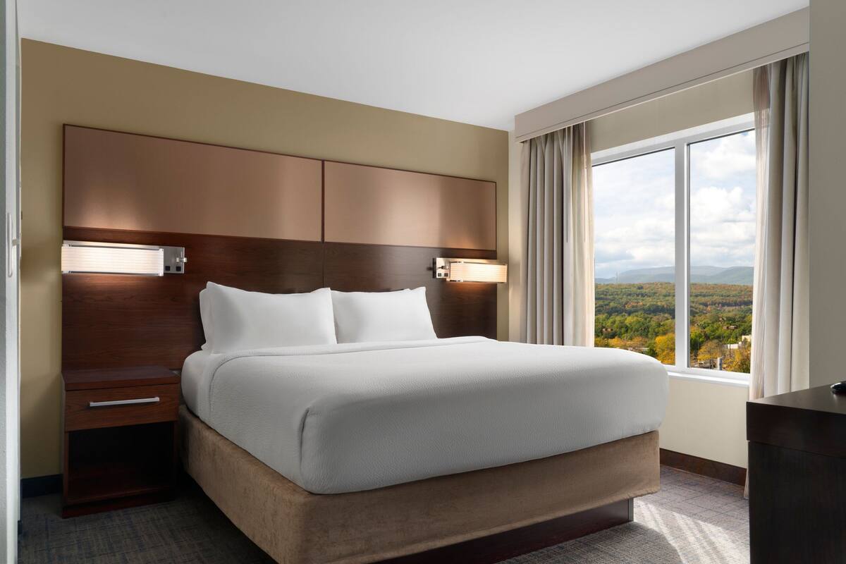 Ein Hotelzimmer mit einem großen Bett, einem Holz-Kopfteil, Nachttischlampen, einem Nachttisch und Blick auf eine gebirgige Landschaft durch ein großes Fenster.