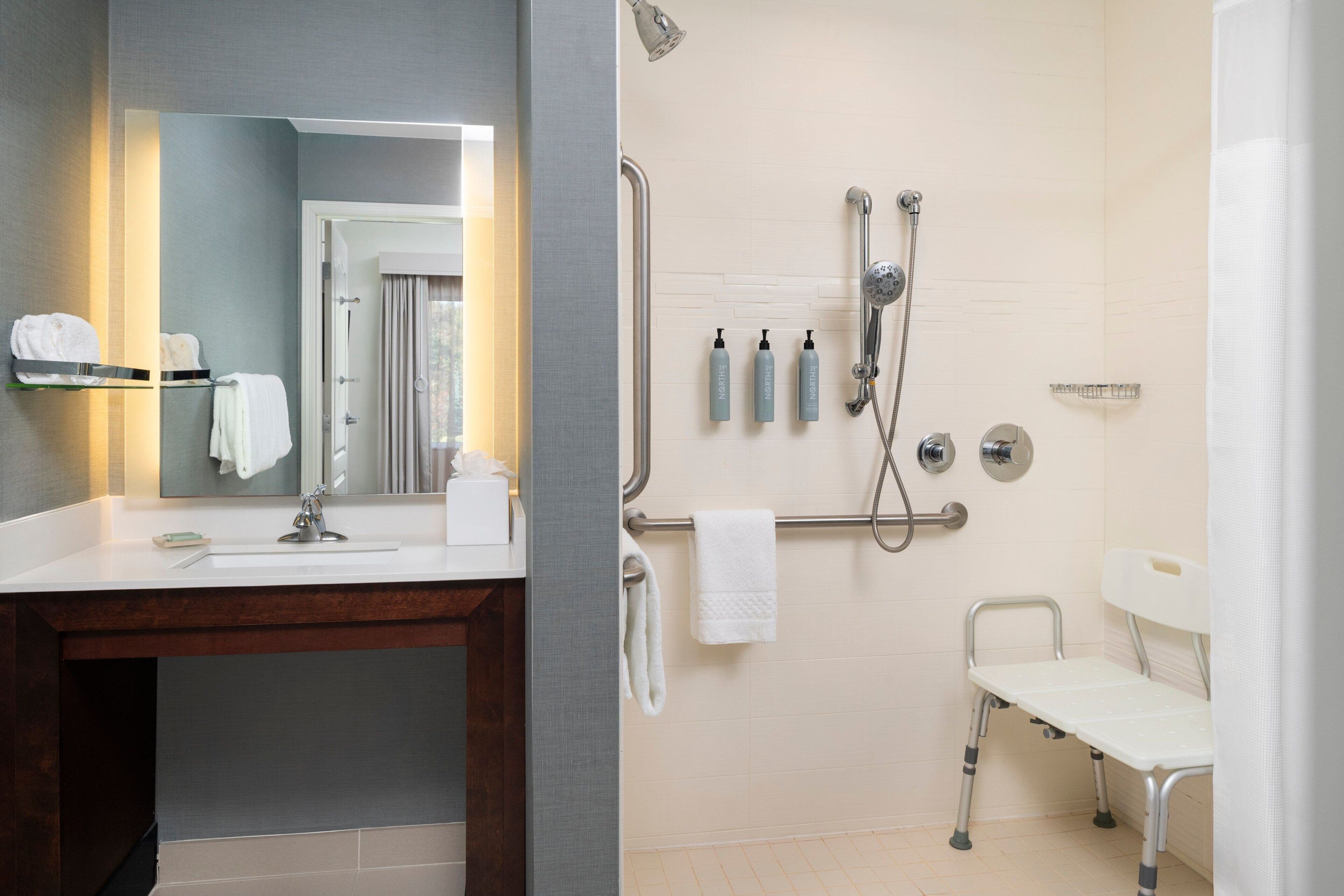 A modern bathroom with a vanity, a shower with grab bars, and a wheelchair-accessible bench.