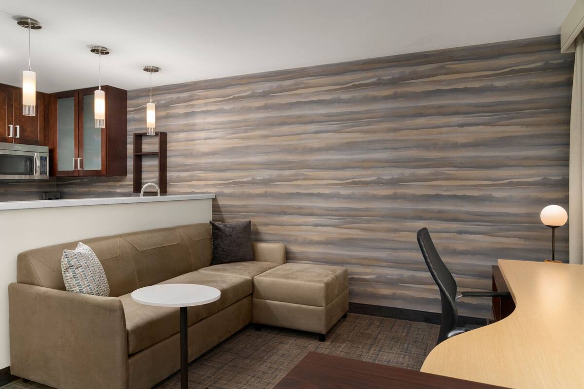 Ein modernes Wohnzimmer mit Ecksofa, einem kleinen runden Tisch und einem Essbereich mit Holztisch und Stuhl. Das Zimmer verfügt über eine Küche mit Mikrowelle und Schränken sowie einer strukturierten Wand.