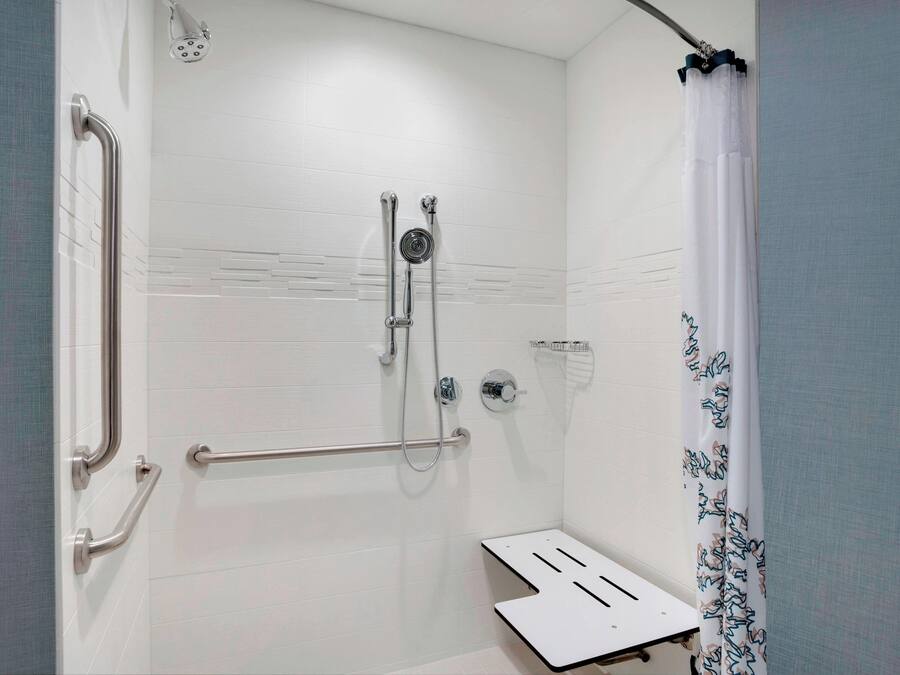 A bathroom with white tiled walls, a shower with a handheld showerhead, a grab bar, and a white shower seat.