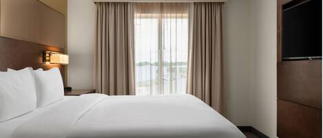 Habitación de hotel con una cama grande, televisión de pantalla plana, mesita de noche con lámpara y ventana con cortinas.