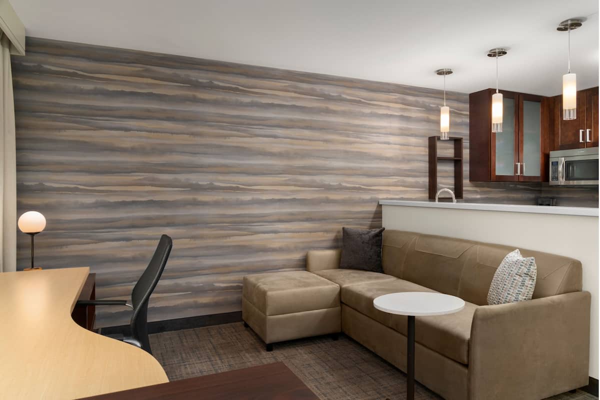 Ein modernes Hotelzimmer mit einer Wand mit Holzoptik, einer Couch, einem kleinen Tisch, einem Sessel und einer Küchenzeile.