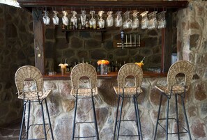 Zona de bar con paredes de piedra, sillas de mimbre y una barra con copas y botellas.