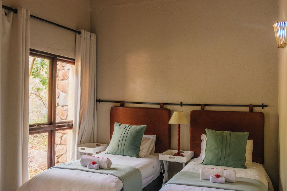 Two single beds with wooden headboards, a window with curtains, and a thatched ceiling.