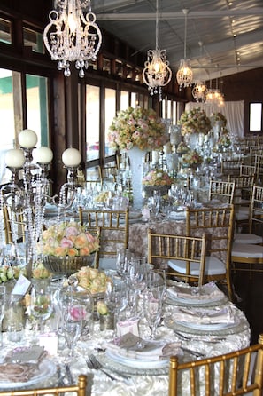 Uma sala de banquetes com longas mesas montadas para um evento formal, decorada com lustres de cristal, arranjos florais e cadeiras douradas.
