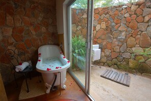 Um banheiro com parede de pedra, uma banheira com pétalas de rosa, uma mesinha com toalha e uma porta de vidro que dá para área externa.