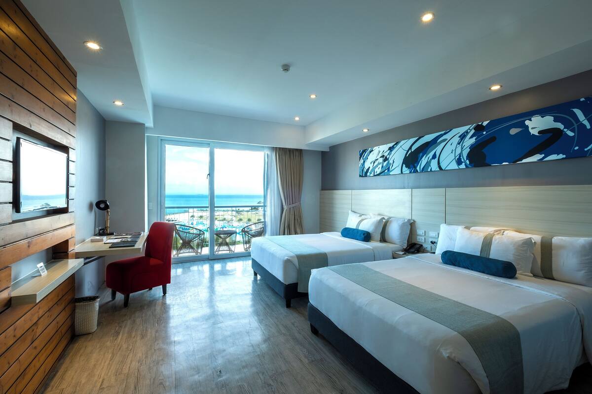 A hotel room with two beds, a desk, a chair, and a large window with a view of the sea.