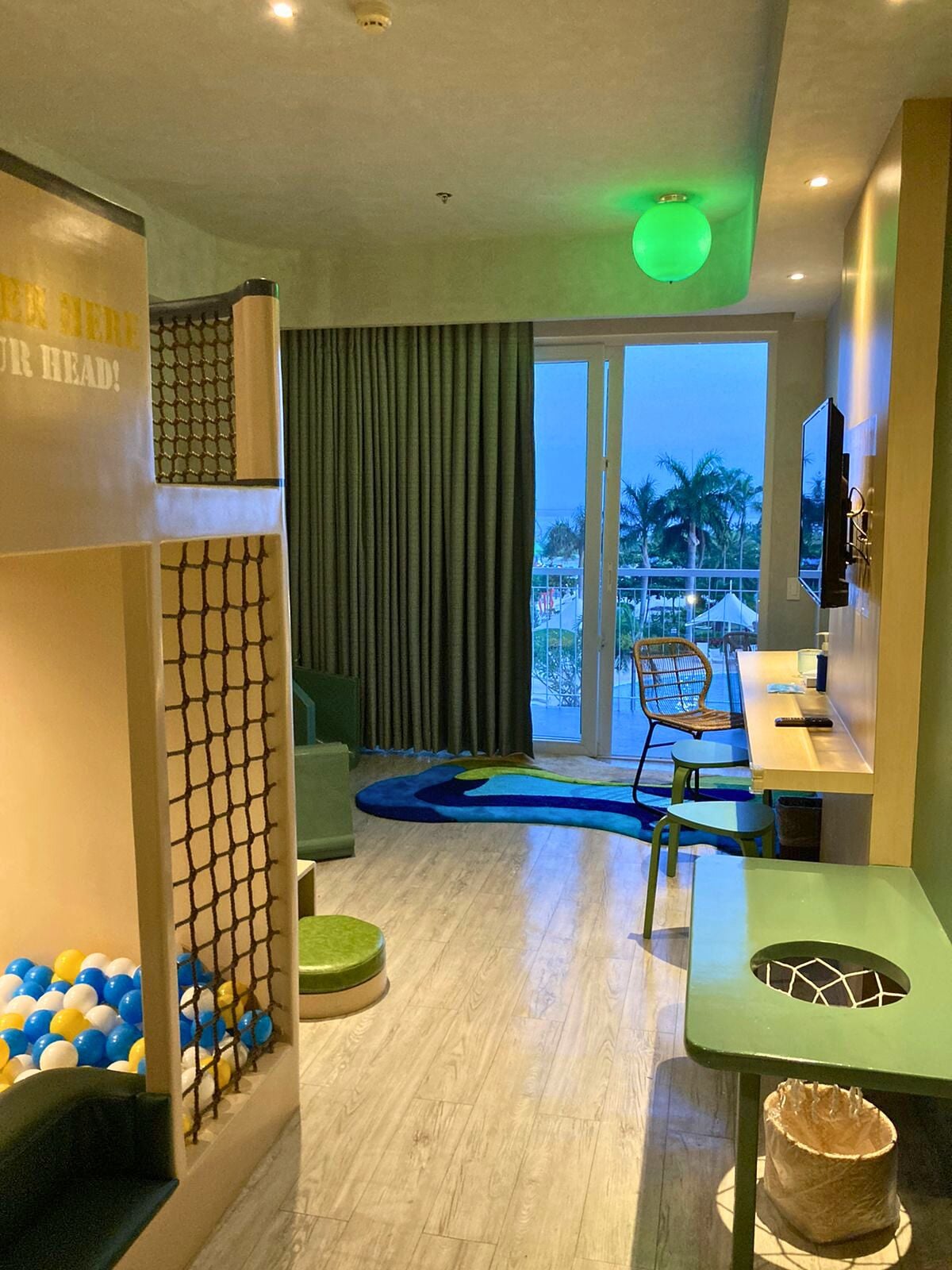 A hotel room with a balcony, a pool area with blue and white balls, a green chair, and a table with a green tablecloth.