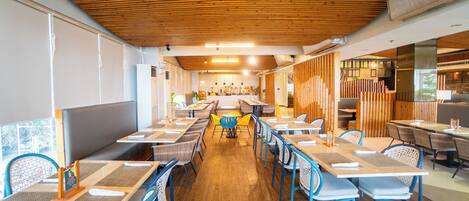 A modern restaurant with wooden ceiling, long tables, and blue chairs.