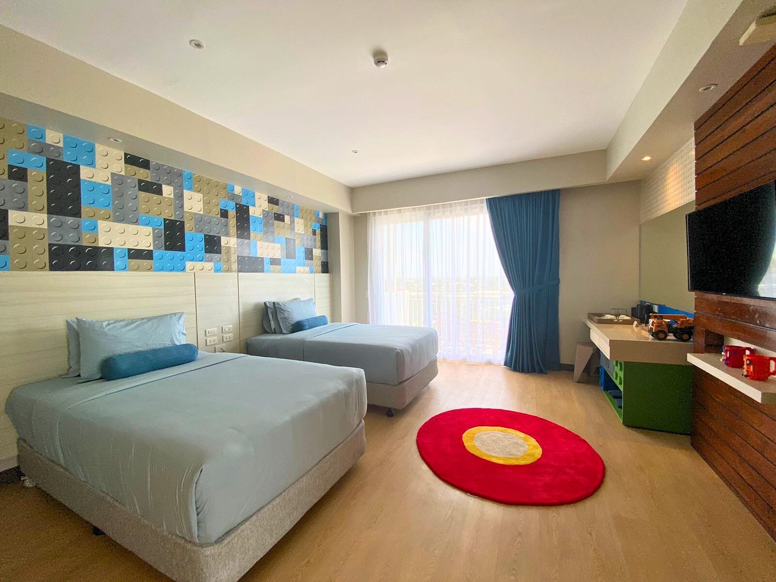 A hotel room with two beds, a TV, a window with curtains, and a wall with a colorful pattern.