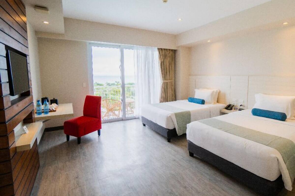 A hotel room with two beds, a TV, a red chair, a small table with water bottles, and a balcony with a view.