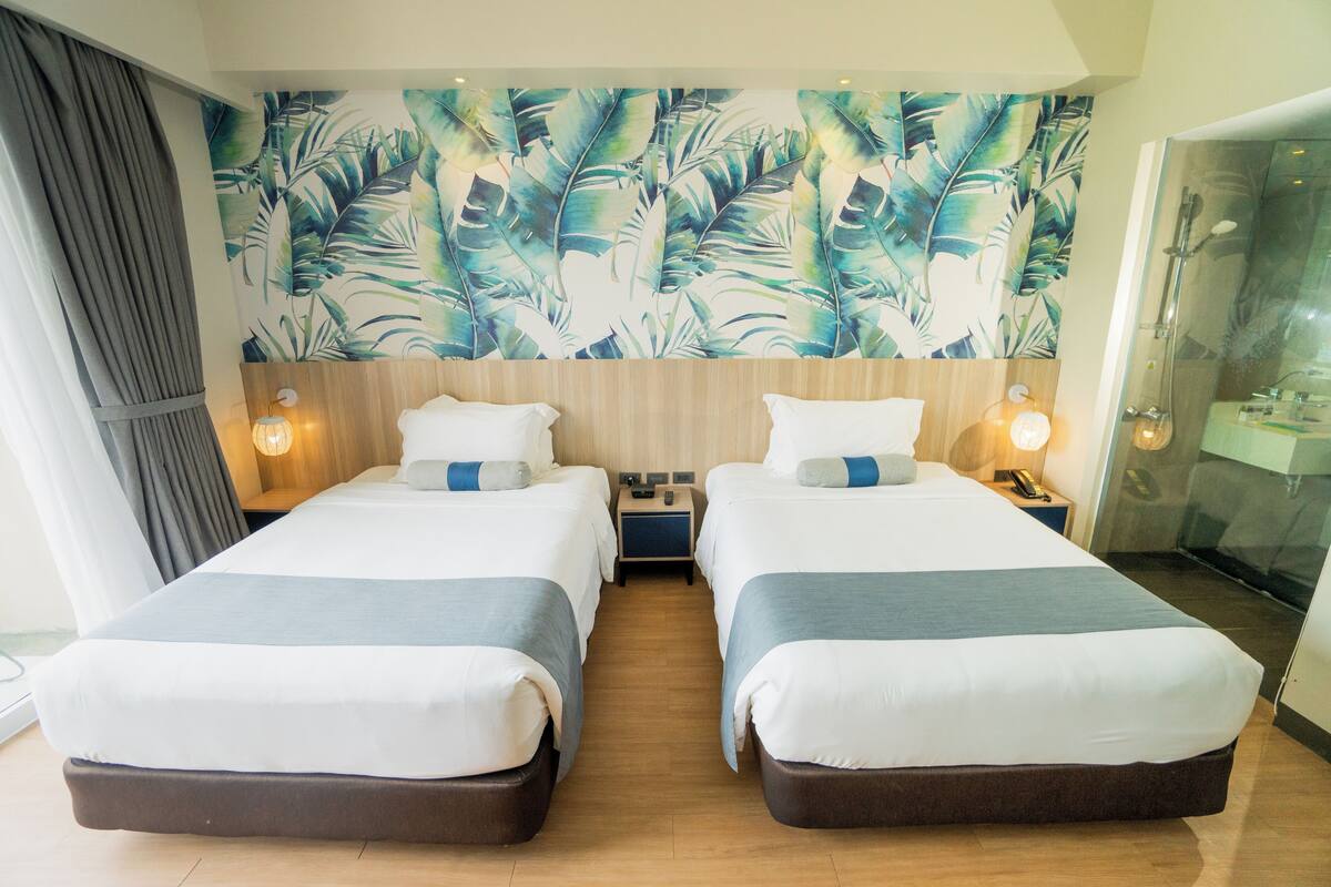 A hotel room with two beds, a large tropical leaf mural, wooden headboard, bedside lamps, and a glass shower enclosure.