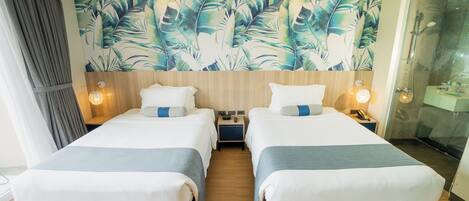 A hotel room with two beds, a large tropical leaf mural, wooden headboard, bedside lamps, and a glass shower enclosure.