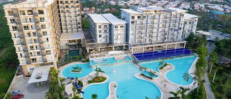 Un hotel de varios pisos con un complejo de piscinas, que incluye una piscina principal y una más pequeña, rodeado de palmeras y vistas de la ciudad al fondo.
