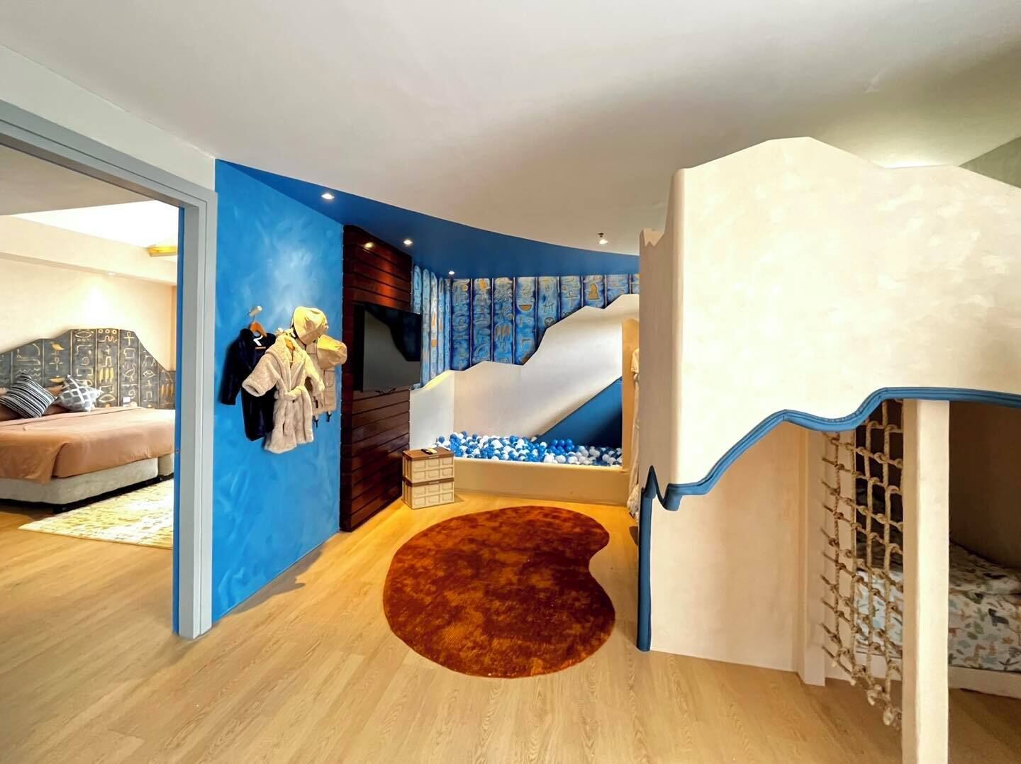 Une pièce avec un mur d’accent bleu, un lit doté d’une tête de lit à motifs, un espace de jeux avec un toboggan et une piscine à balles, et un tapis rond marron.