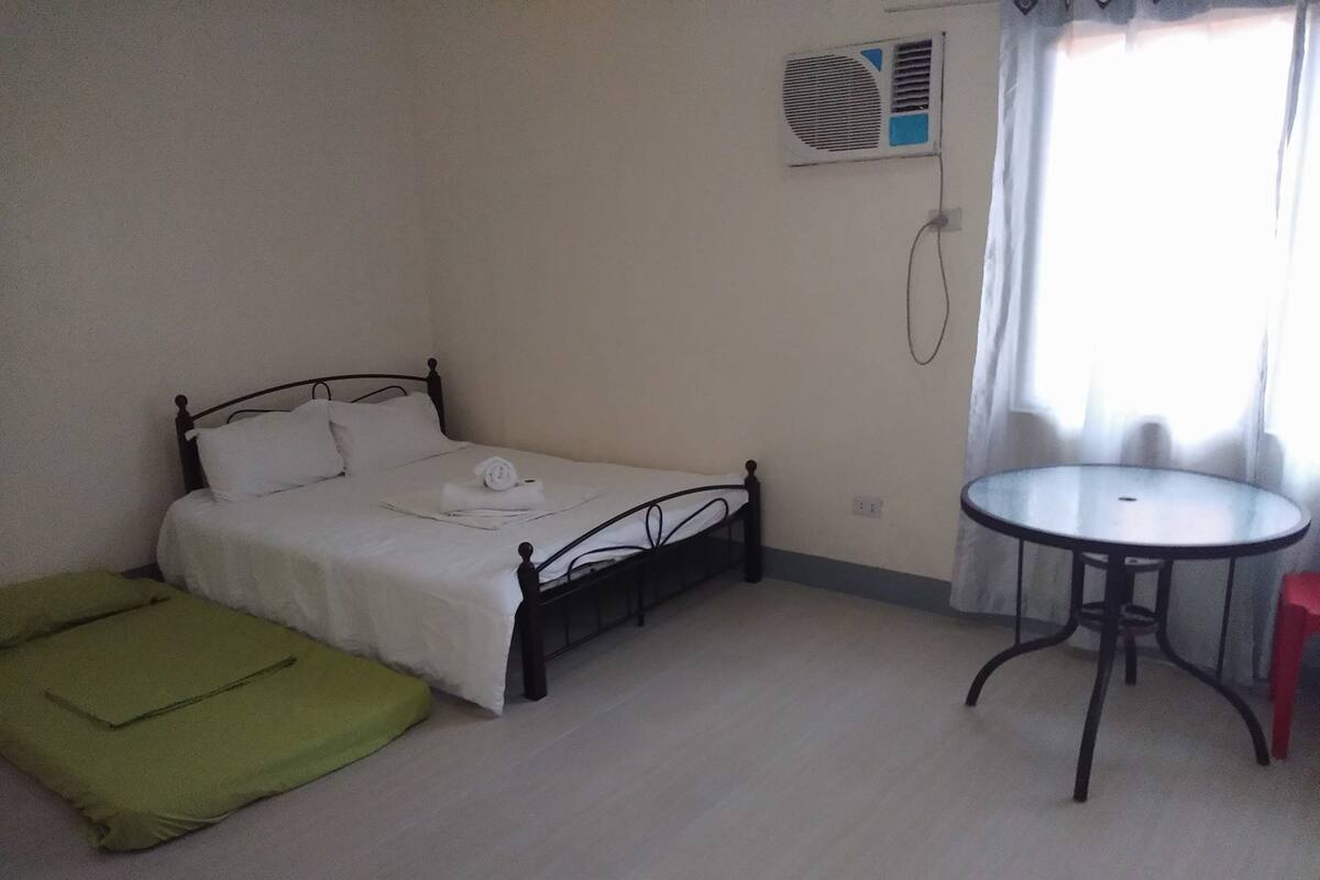 Une petite pièce propre, avec un lit simple, un coussin vert, une petite table ronde et une fenêtre avec des rideaux.