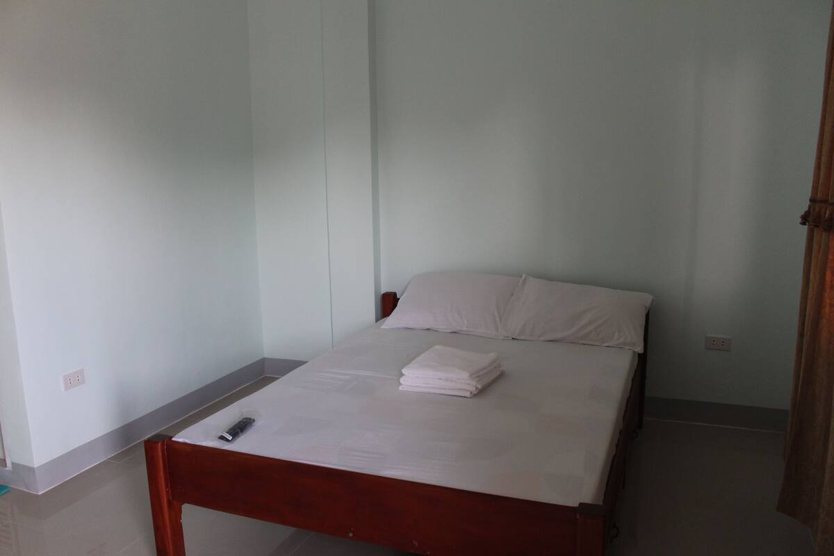 Une chambre simple et dépouillée, avec un lit simple, une tête de lit en bois et une télécommande posée sur le lit.