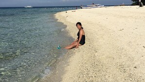 En sandstrand med klart vatten, en båt i fjärran och en person som sitter på stranden.