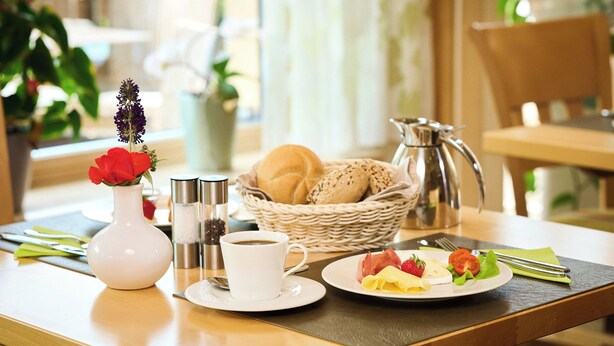 Una escena de desayuno con un jarrón blanco lleno de flores rojas, una taza de café, un plato de comida y una cesta de pan.